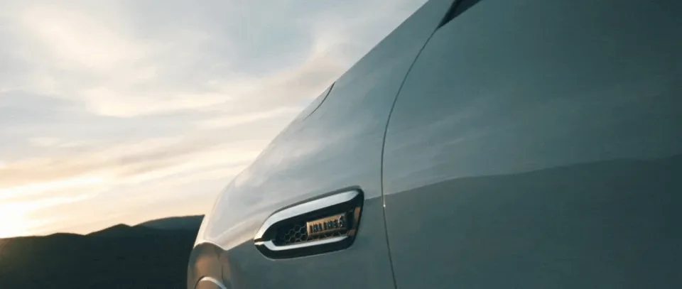 Nahaufnahme der Frontpartie eines modernen Fahrzeugs mit LED-Scheinwerfer vor bewölktem Himmel und Bergsilhouette im Hintergrund bei Dämmerung.