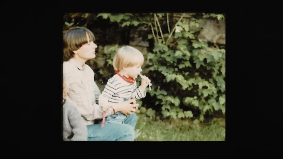 Zwei Kinder im Garten, ein älteres Kind mit braunem Haar und ein jüngeres blondes Kind in gestreiftem Pullover, vor grünem Laubwerk im Hintergrund.