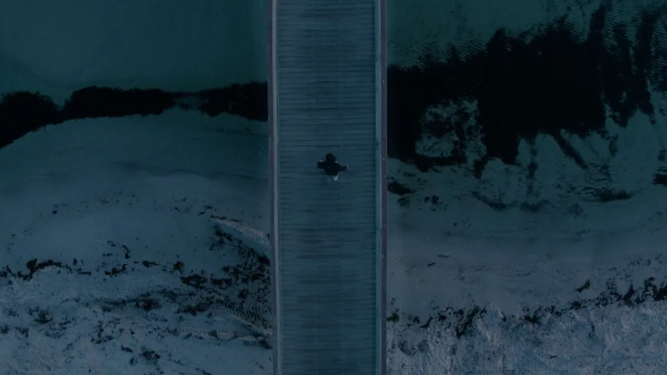 Luftaufnahme einer Person, die allein auf einem Holzsteg über ruhigem Wasser sitzt, umgeben von verschneitem Ufer und Winterlandschaft.
