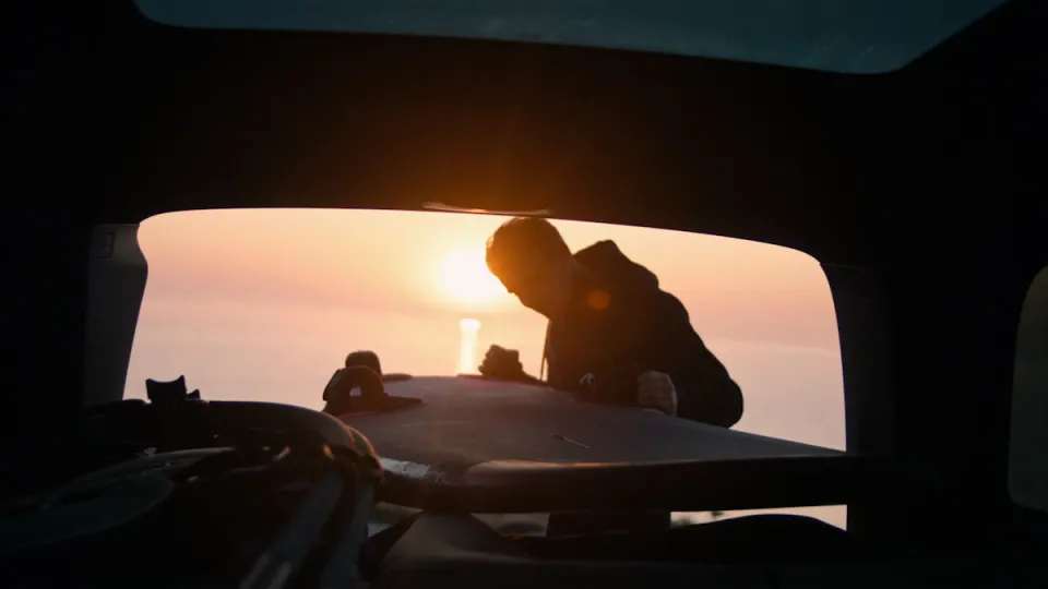 Silhouette einer Person, die bei Sonnenaufgang aus dem geöffneten Kofferraum eines Autos blickt, mit Meer im Hintergrund.