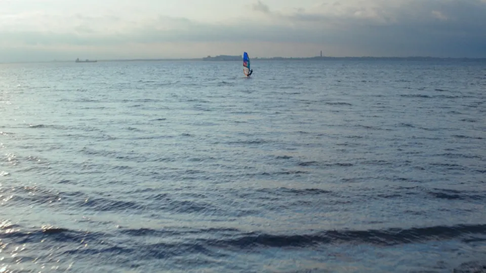 Windsurfer mit blauem Segel auf ruhigem Meer bei bewölktem Himmel, entfernte Küstenlinie im Hintergrund.