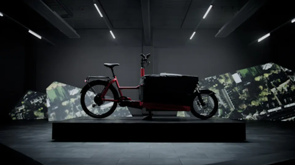 Rotes Lastenfahrrad auf schwarzem Podest in minimalistischem Ausstellungsraum mit Tarnmuster-Objekten im Hintergrund