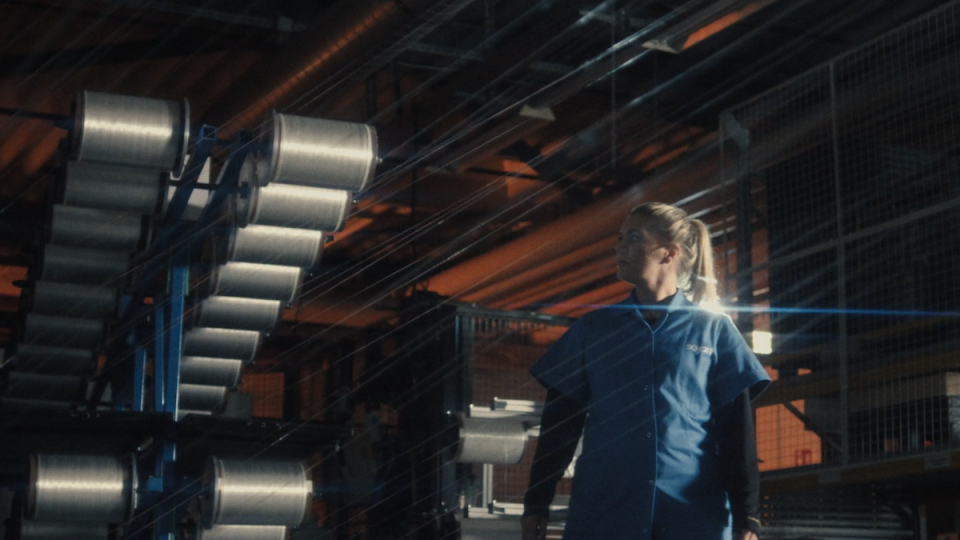 Fabrikarbeiterin in blauer Arbeitskleidung steht vor einem Lagerregal mit Metallrollen in einer Industriehalle.