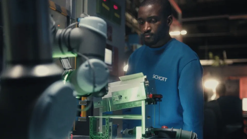 Mann in blauem Schott-Pullover arbeitet mit Roboterarm an technischer Maschine in einer Werkstatt.
