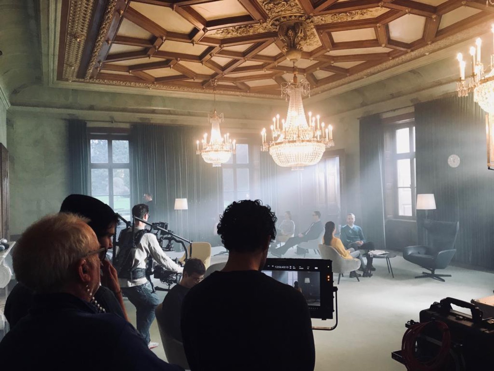 Filmteam bei Dreharbeiten in einem eleganten Raum mit Kassettendecke und Kristallkronleuchtern, während Schauspieler auf Möbeln im Hintergrund posieren.