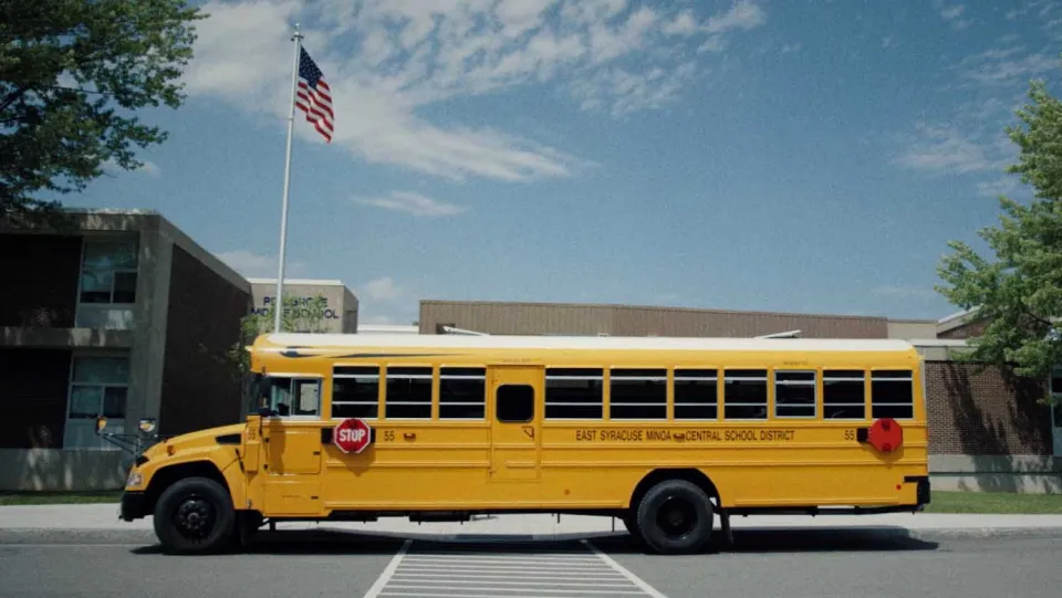 Gelber Schulbus des East Syracuse Minoa Central School District parkt vor einem Schulgebäude mit US-Flagge