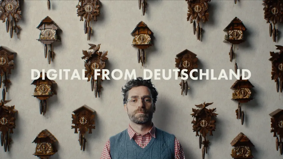Mann mit Brille und Pullunder vor Wand mit vielen Kuckucksuhren, darüber der Text "DIGITAL FROM DEUTSCHLAND"