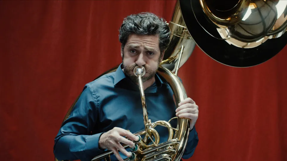 Mann mit dunklem Haar und Bart spielt Tuba vor rotem Vorhang, trägt blaues Hemd.