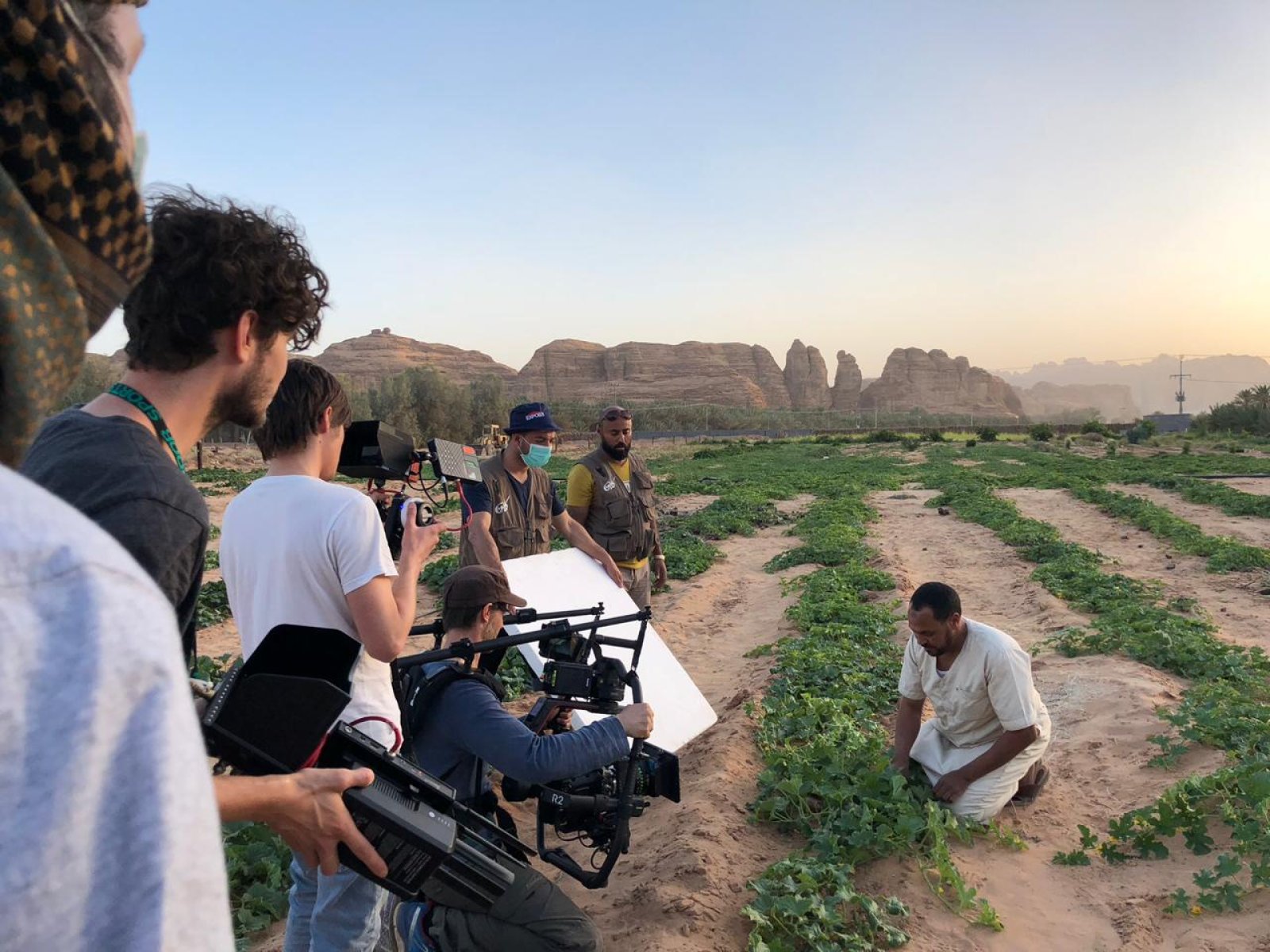 Filmteam bei Dreharbeiten auf einem Feld mit Kameramännern, Crew-Mitgliedern und einem knienden Darsteller, im Hintergrund Felsformationen bei Sonnenuntergang.